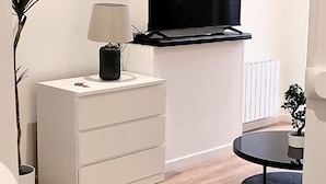A modern living room with a flat-screen TV mounted on the wall, a white side table with a lamp, a round coffee table, and a potted plant. - Le Cantin 4 Pers Wifi Courette Bollaert (Lens)