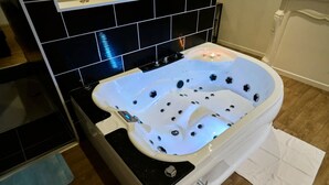 A bathroom with a white bathtub, black tiled walls, and a wooden floor. - Baignoire Blaneo Sauna 10 Minutes de la Gare (Tourcoing)