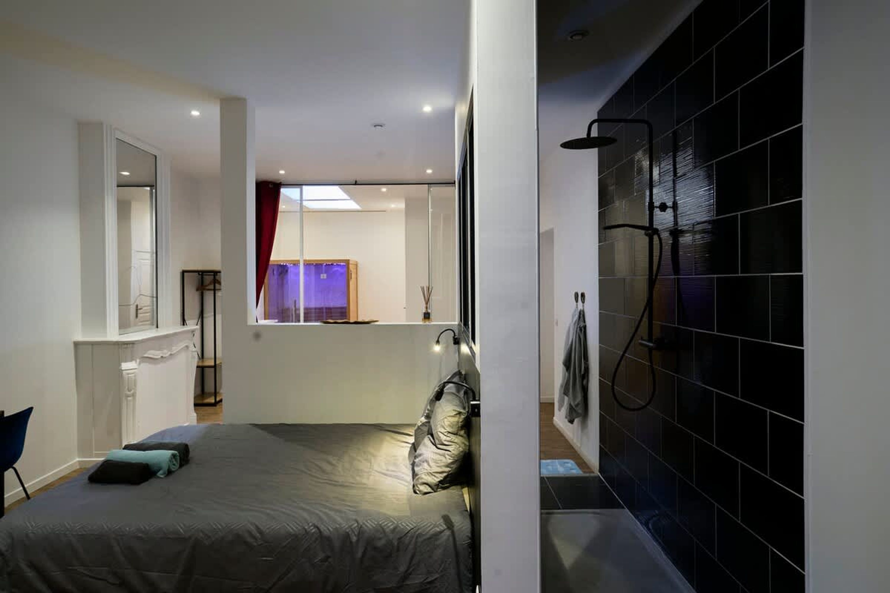 A compact hotel room with a bed, a small desk, a mirror, a shower area, and a view of a cityscape.