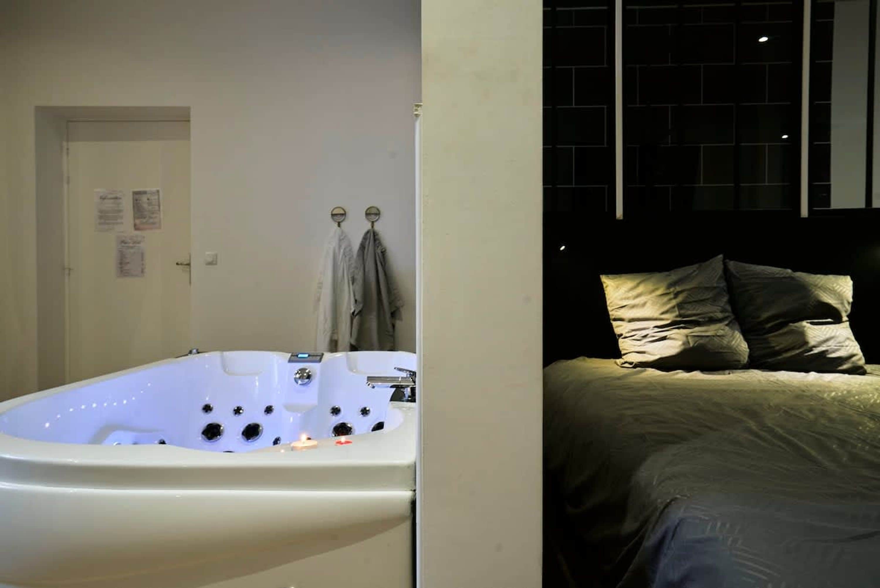 A room with a bathtub, a bed with pillows, a door with a towel rack, and a window with a brick pattern.
