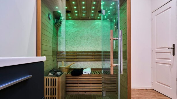 A glass-enclosed sauna with wooden benches, a wooden floor, and a white door. - Baignoire Blaneo Sauna 10 Minutes de la Gare (Tourcoing)