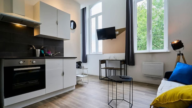 Una cocina moderna con horno integrado, comedor con mesa y sillas, televisor de pantalla plana montado en la pared y una cama con almohada amarilla.