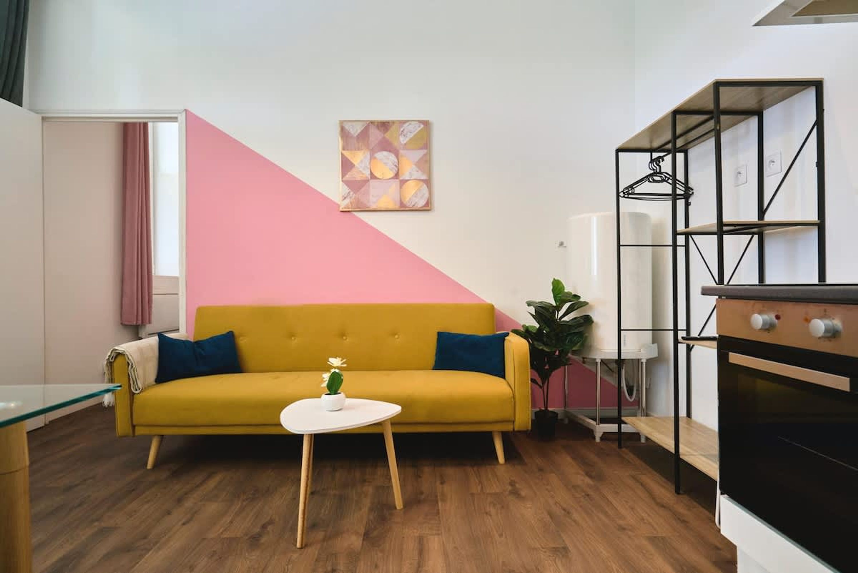 Un salon moderne avec un canapé jaune, une petite table blanche, un coin cuisine doté d’un four noir et un mur décoré de motifs roses et blancs.