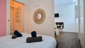 Ein Schlafzimmer mit einem Bett, einem an der Wand befestigten Spiegel, einem Nachttisch und Blick auf ein Badezimmer mit Handtuchhalter.