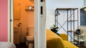 Ein kompakter Wohnraum mit Bett, gelber Couch, kleinem Schreibtisch und einem Badezimmer mit Toilette und Waschbecken.