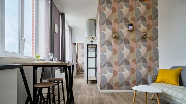 A modern kitchen with a bar area, a dining table with stools, a sofa with a yellow pillow, and a patterned wall. - En D Placement 5 Calme et Lumineux (Tourcoing)