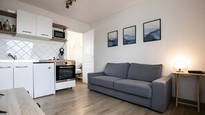 Moderni keittiö, jossa on valkoiset kaapit, mikroaaltouuni ja uuni. Olohuoneessa on harmaa sohva ja pieni pöytä, jossa on lamppu.