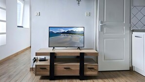 TV-taso on valmistettu puusta, ja sen päällä on televisio. Taustalla näkyy rantamaisema, valkoinen ovi ja ikkuna, jossa on sälekaihtimet.
