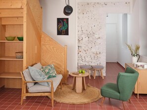 Un salon chaleureux avec un escalier en bois, une œuvre d’art représentant un coq aux couleurs vives et un mur en briques rustiques.