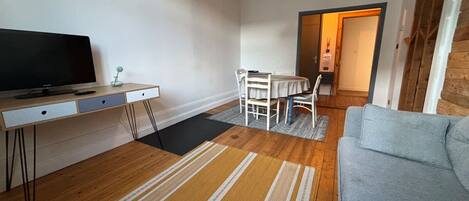 A modern living room with a flat-screen TV, a dining table with chairs, a grey sofa, and a wooden floor.