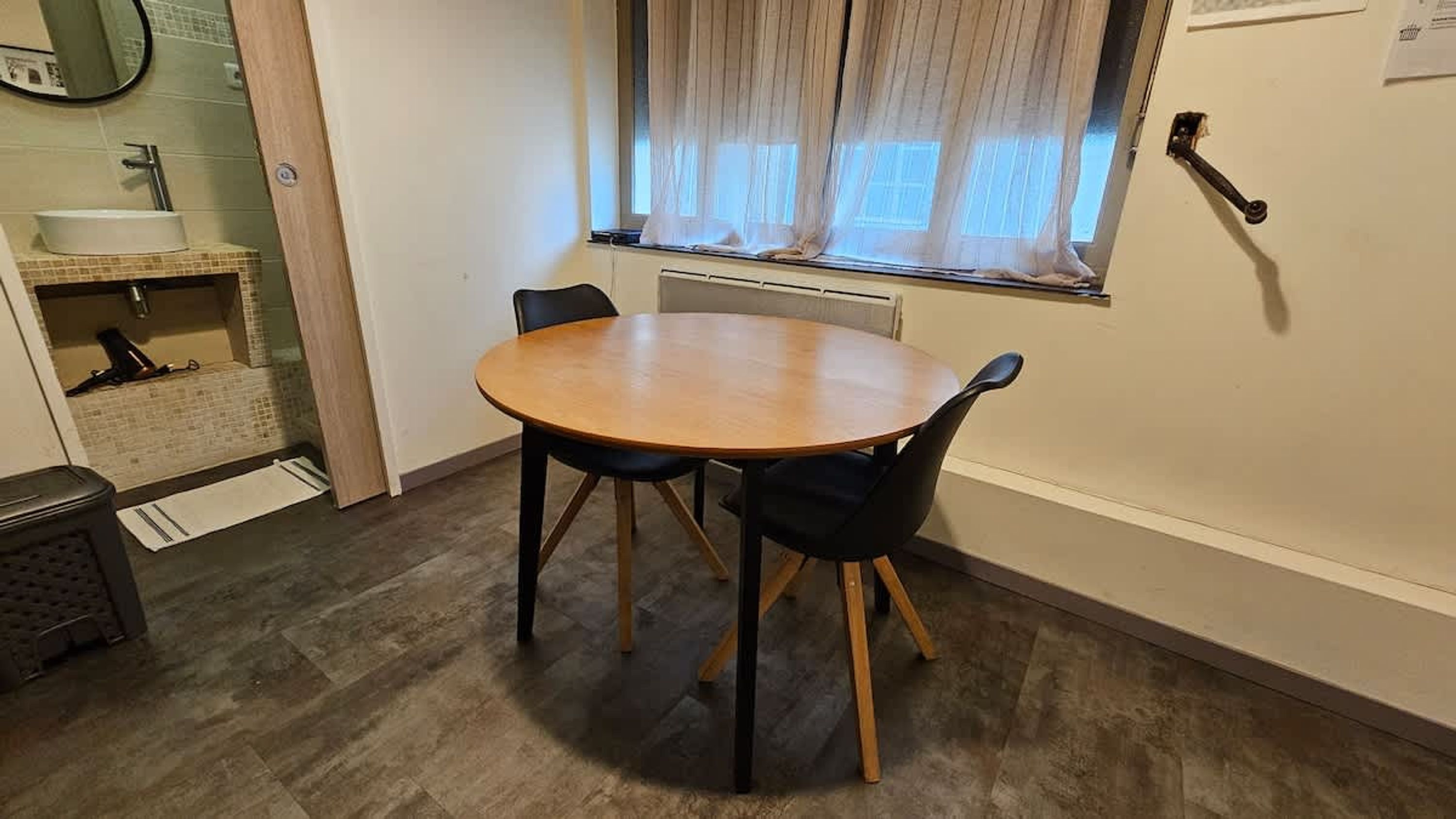 A small dining area with a round table and two chairs, a window with sheer curtains, a radiator, and a sink with a mirror above it.