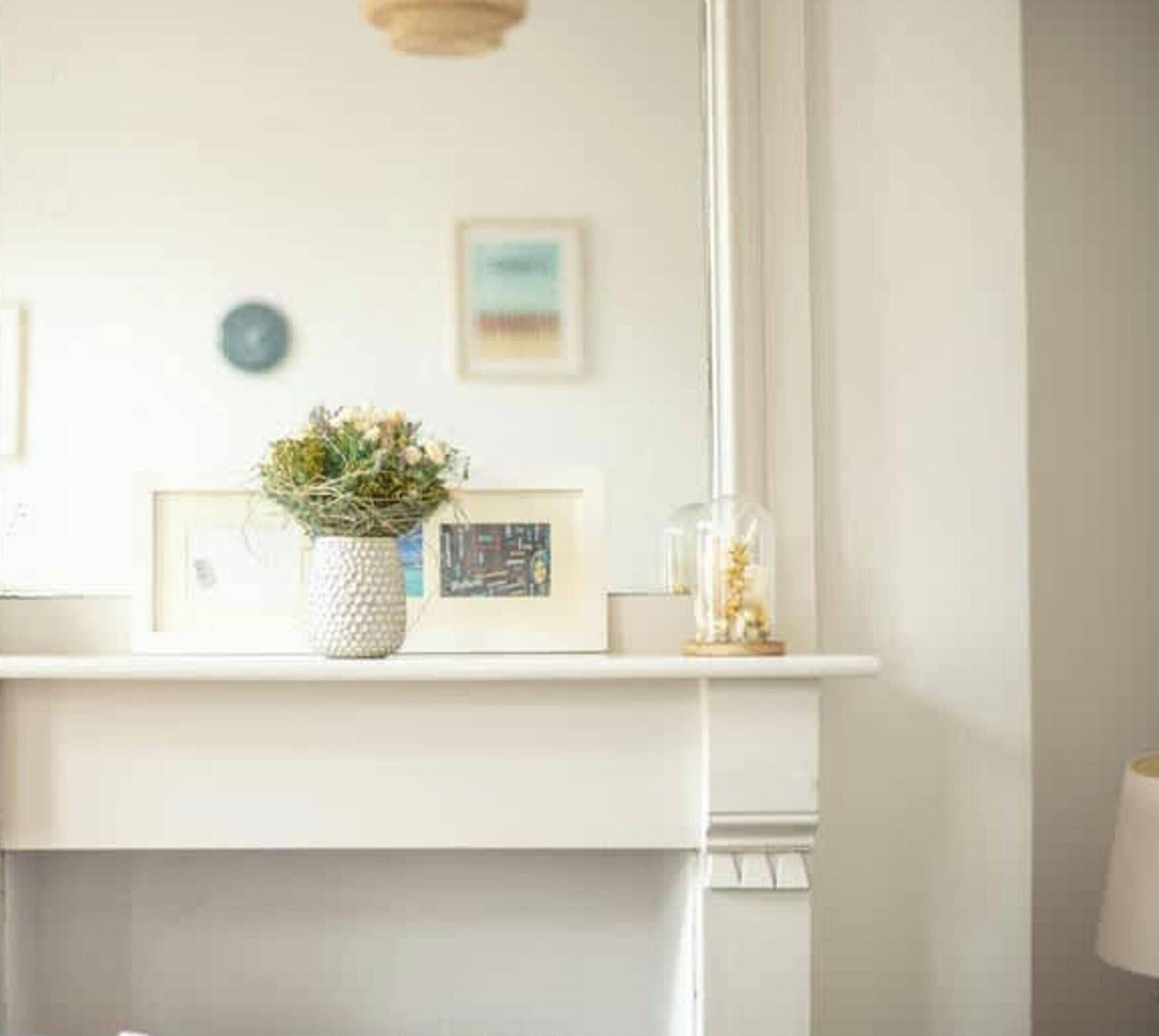 A white mantelpiece with a framed picture, a vase with flowers, and a glass jar with a plant.
