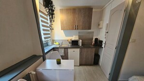 A compact kitchenette with a microwave, coffee maker, and a small refrigerator. There is a white table with a chair, and a window with blinds. - Le Studio du Th tre (Arras)