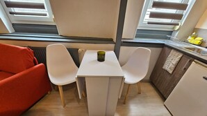 A compact room with a white table, two white chairs, a red sofa, a small kitchenette, and a window with blinds. - Le Studio du Th tre (Arras)