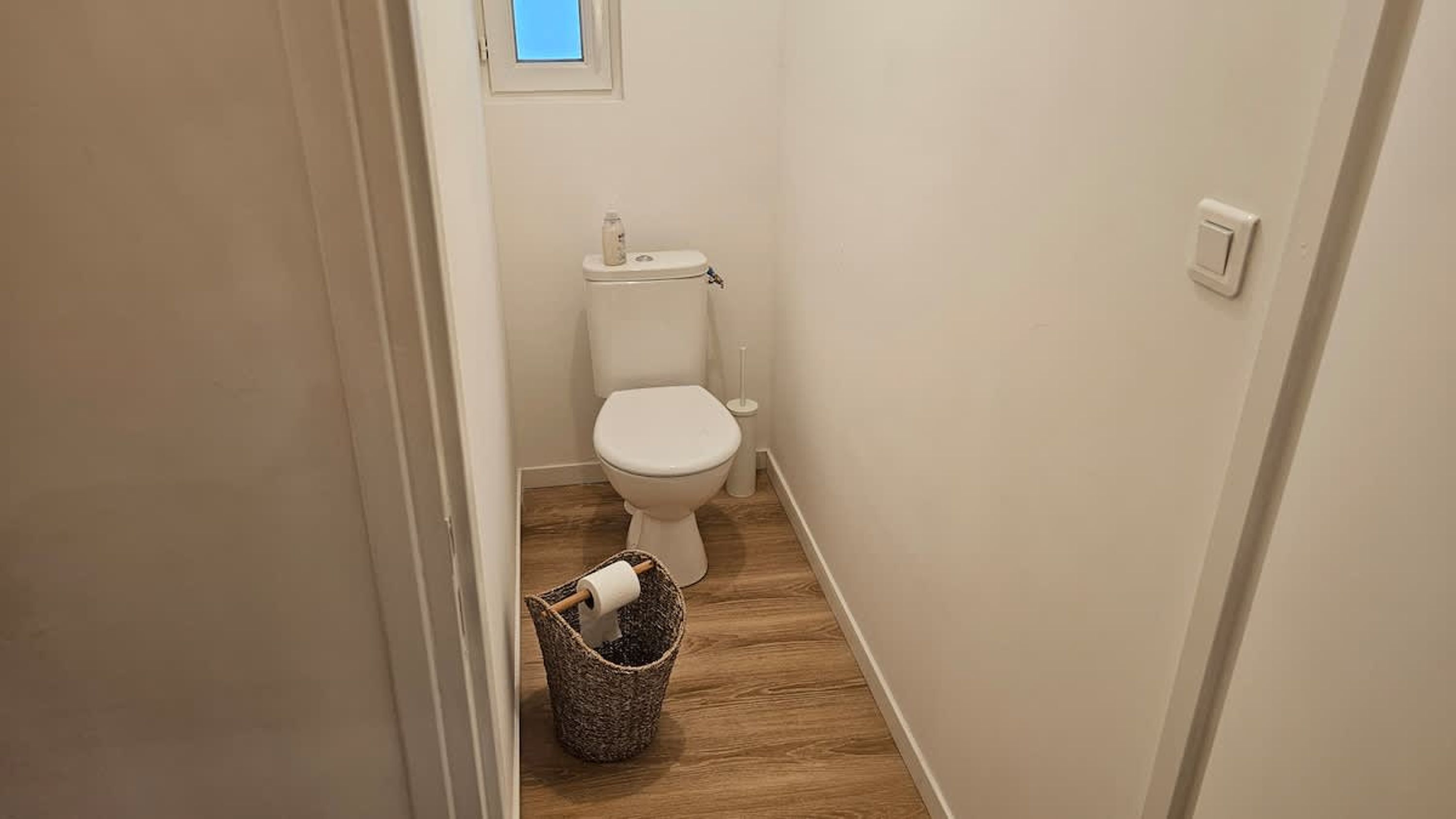 A small, narrow bathroom with a white toilet, a wooden floor, and a trash bin.