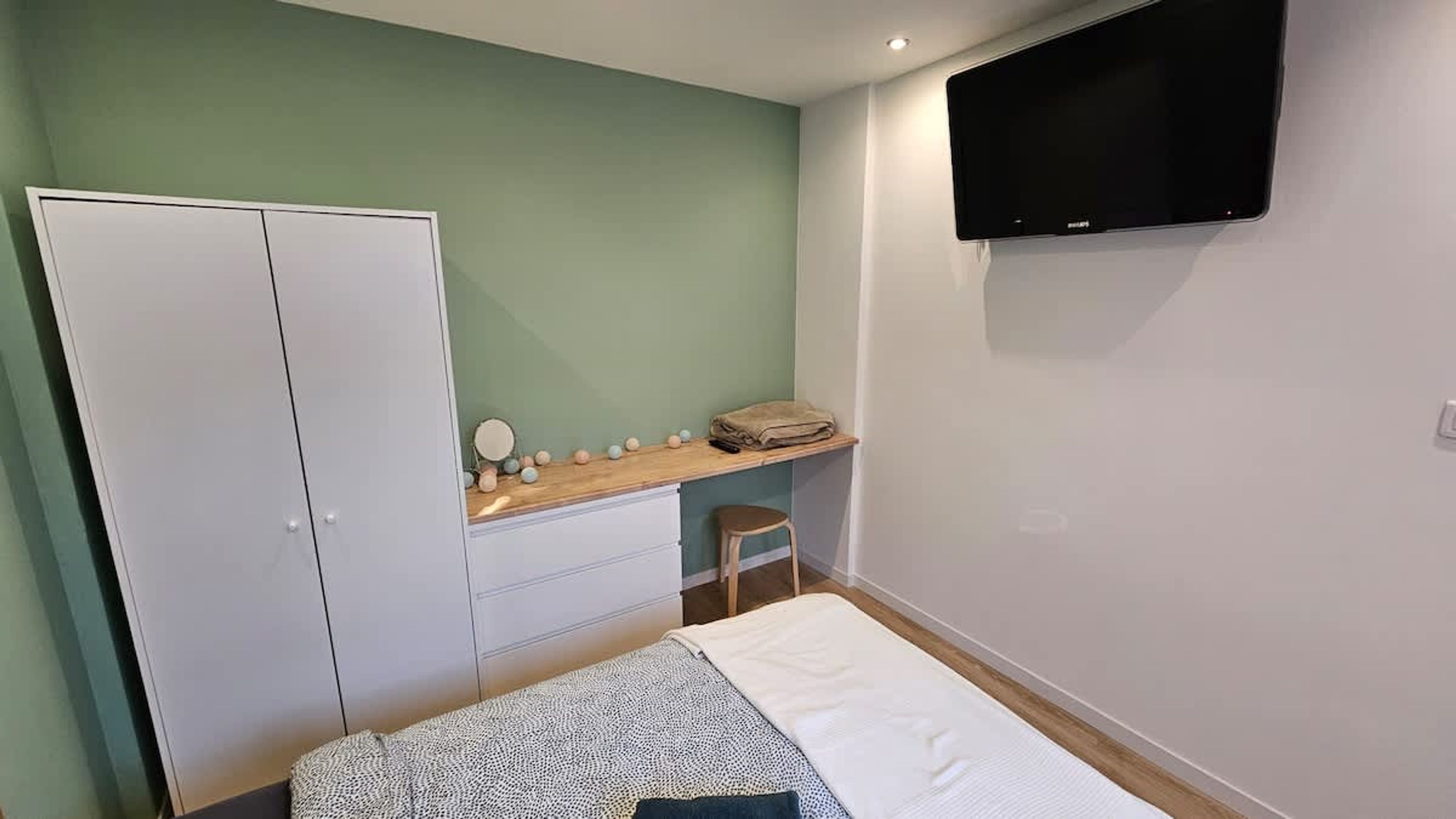A bedroom with a bed, a wardrobe, a vanity with a mirror, a shelf with toiletries, and a TV mounted on the wall.