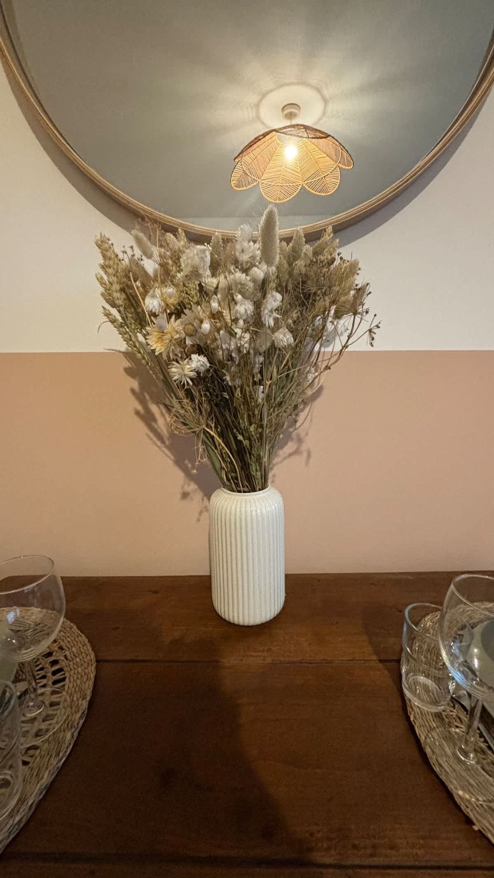 Een witte vaas met gedroogde bloemen op een houten tafel, een spiegel aan de muur en een hanglamp.