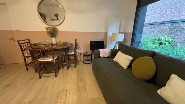 A living room with a dining area, a round table, wooden chairs, a sofa, a mirror, and a window with a view of greenery. - Le Citadin Appartement Arras Centre 6 Personnes (Arras)