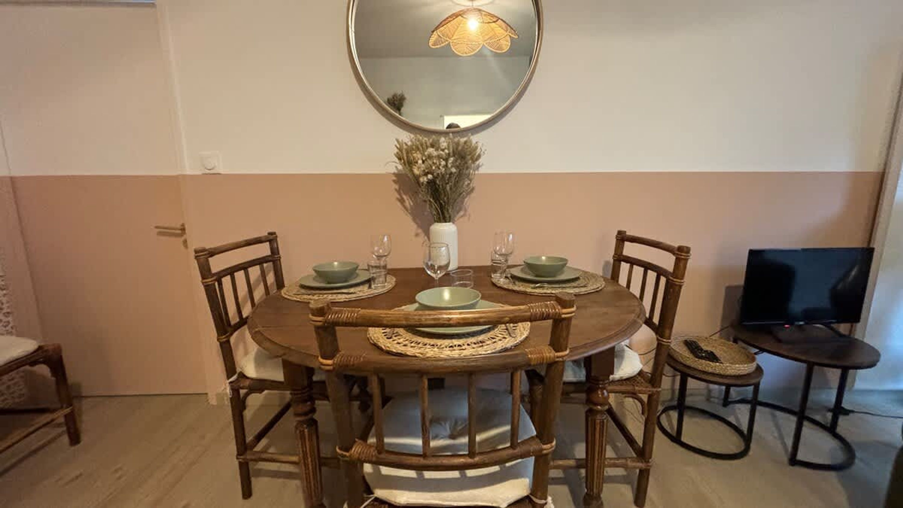 Una mesa de comedor con cuatro sillas, un jarrón con flores secas, un televisor y un espejo en la pared.