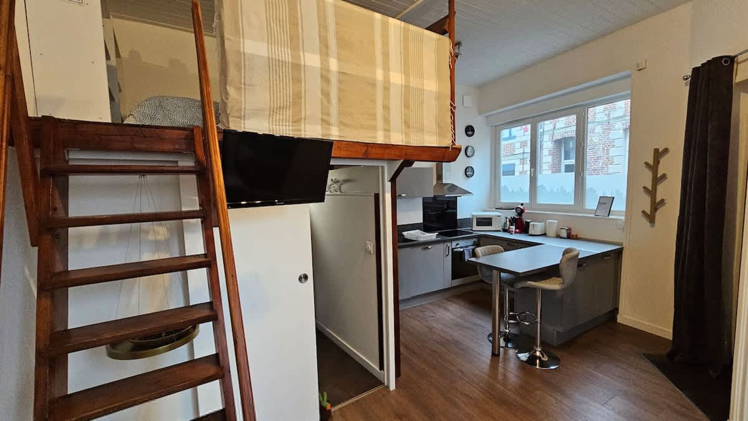 A compact kitchen with a refrigerator, microwave, and stove. A wooden ladder is against the wall, and a small table with a chair is near the window.