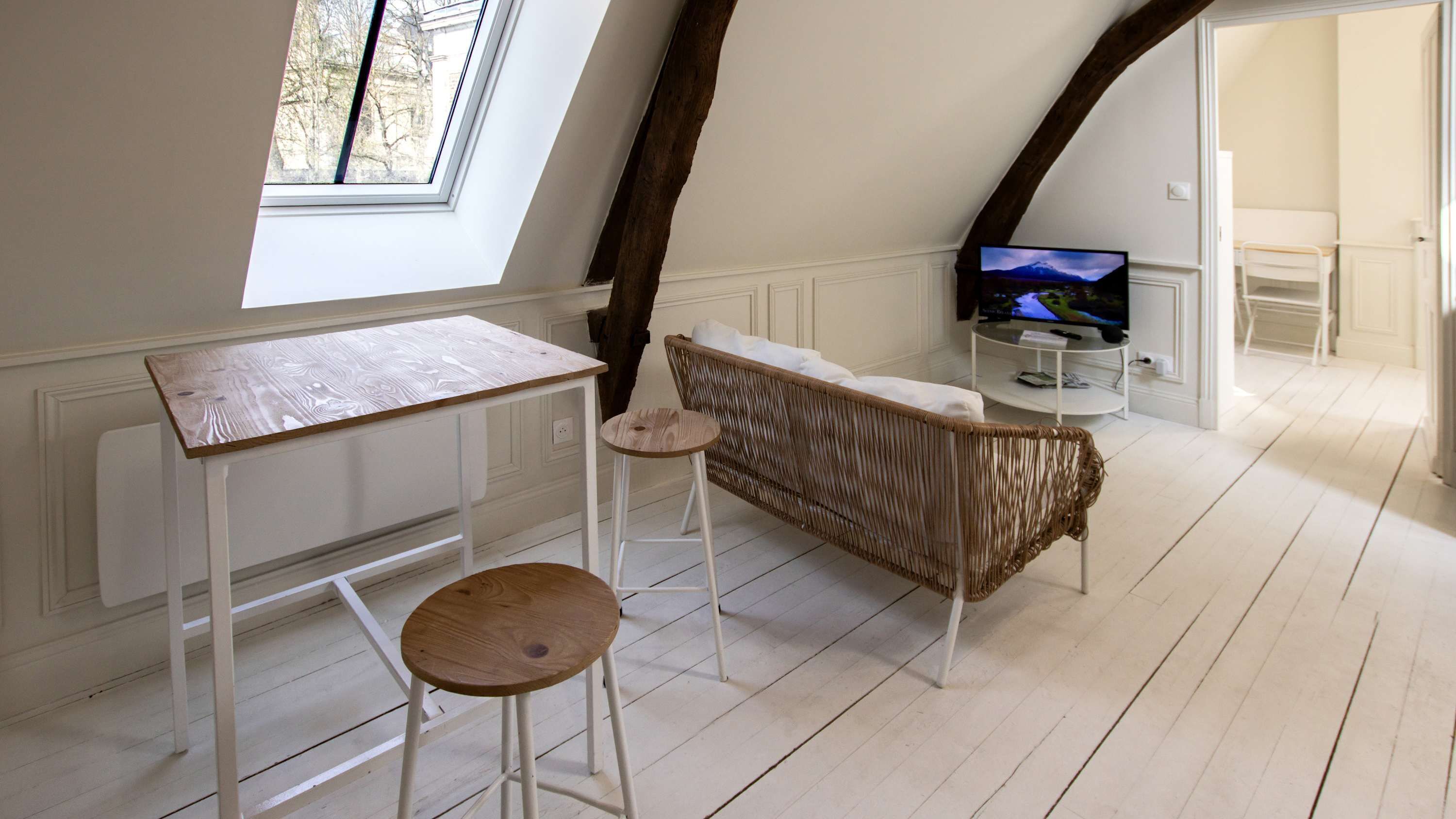 A room with a sloped ceiling, a television on a stand, a small table with stools, and a wicker sofa.