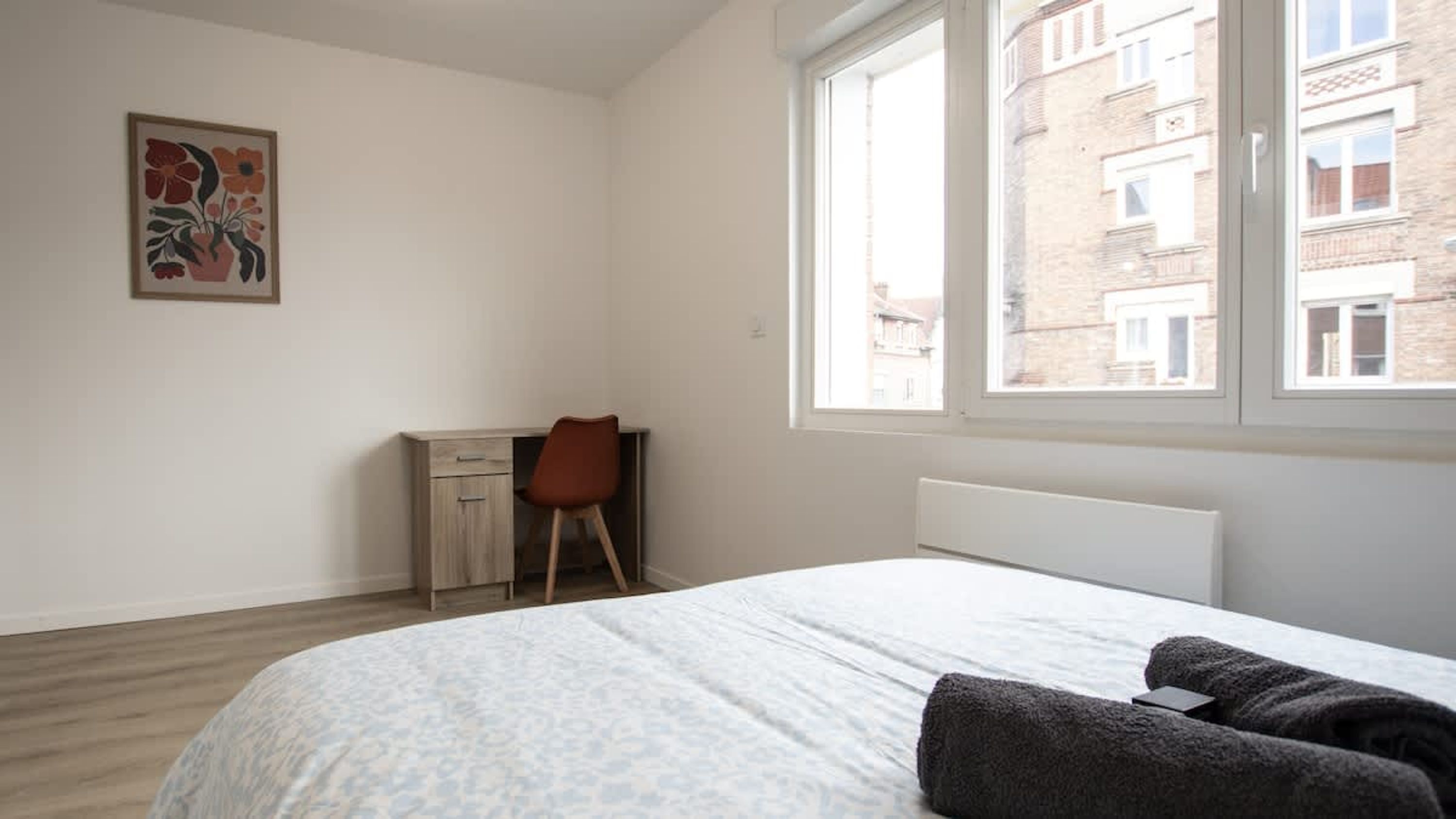 A bedroom with a bed, a desk, a chair, a radiator, and a window with a view of buildings.