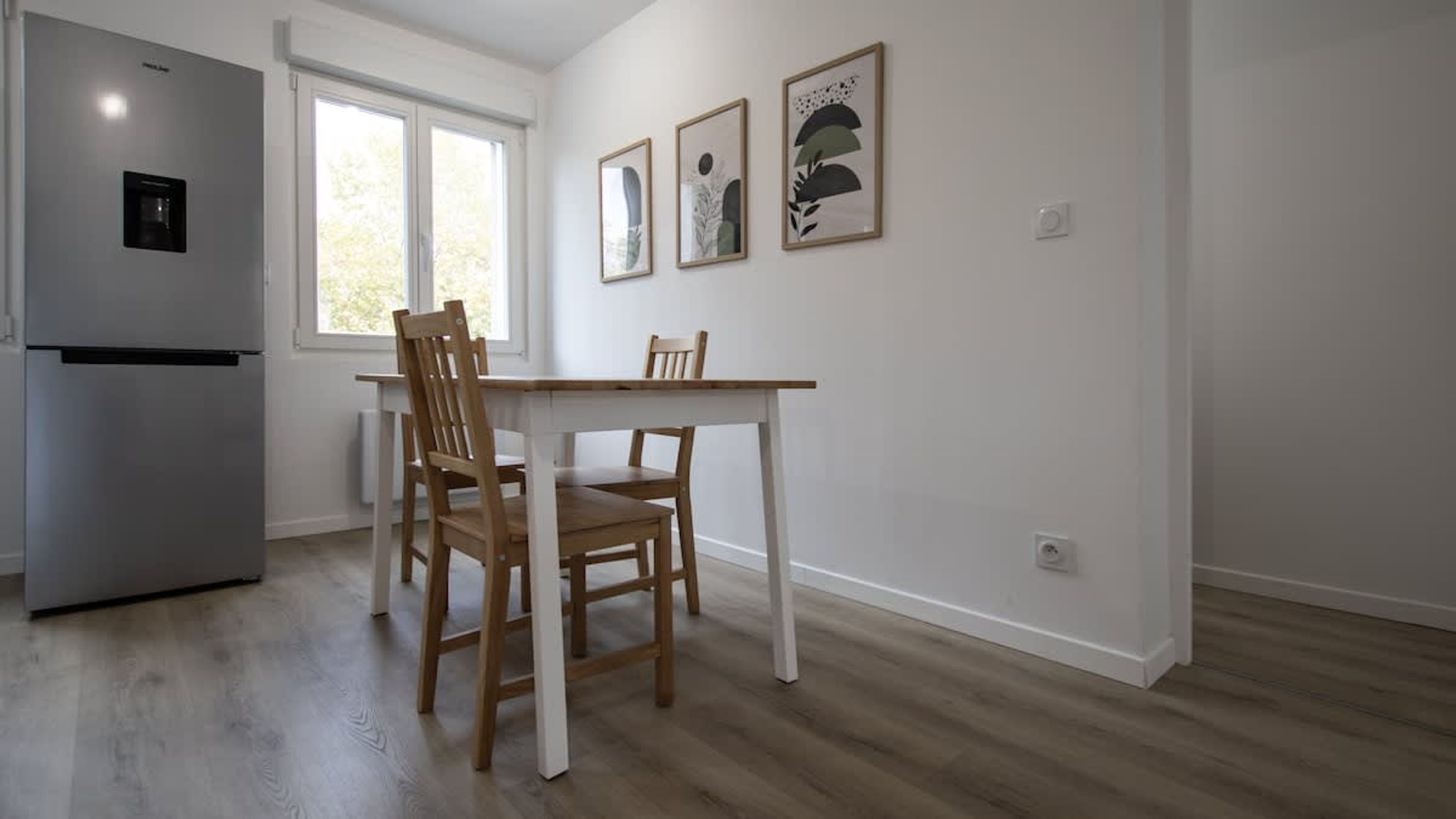 A kitchen with a refrigerator, a dining table, and two chairs. There are three framed artworks on the wall.