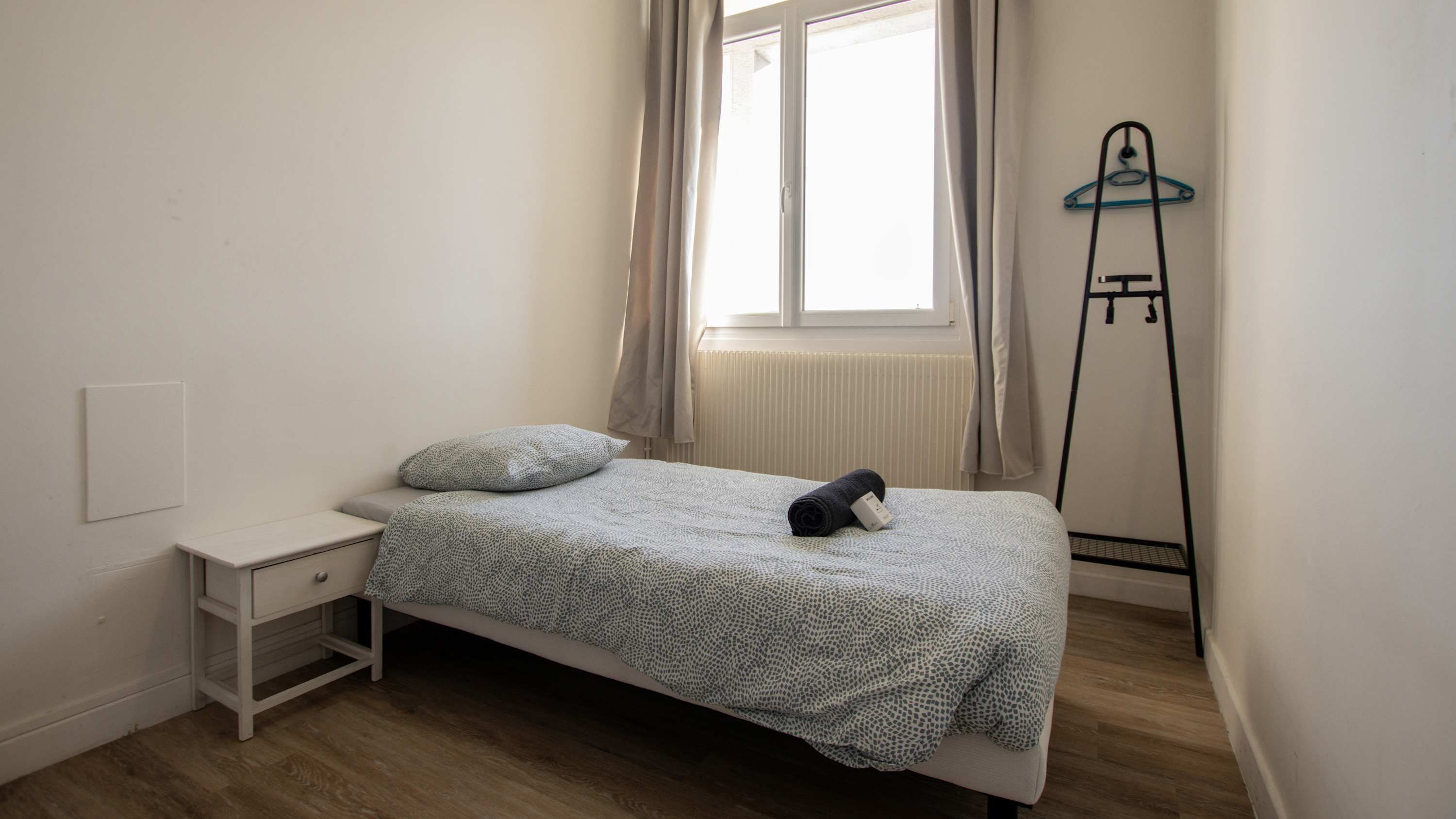 A small, single-bed room with a window, a radiator, a bedside table, and a coat rack.