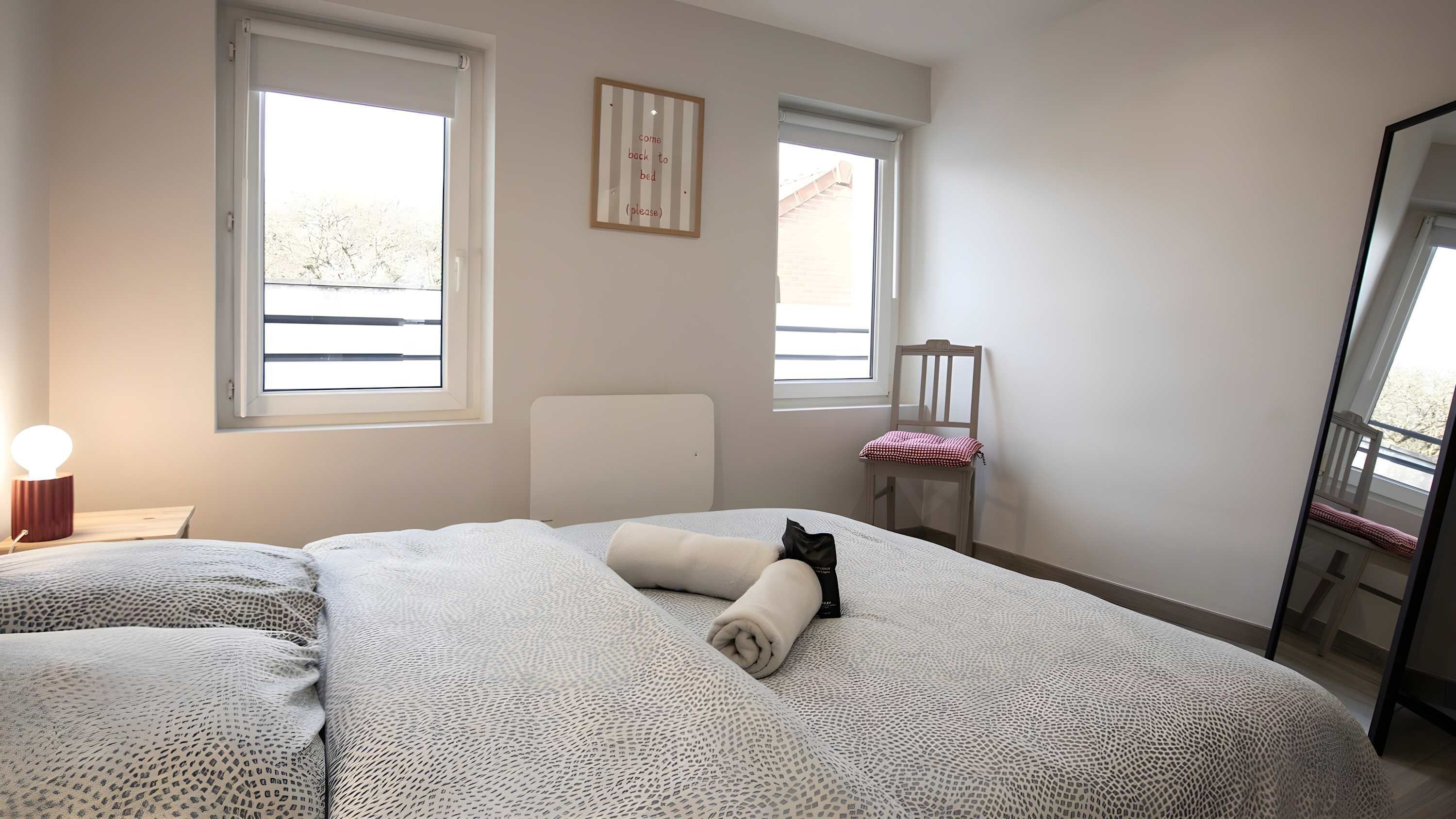A bedroom with two beds, a bedside table with a lamp, a chair, and a window with a view of trees.