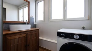 A modern bathroom with a wooden vanity, a white sink, a mirror, and a front-loading washing machine. - Le Perchoir de Maurice Calme et Cosy (Arras)