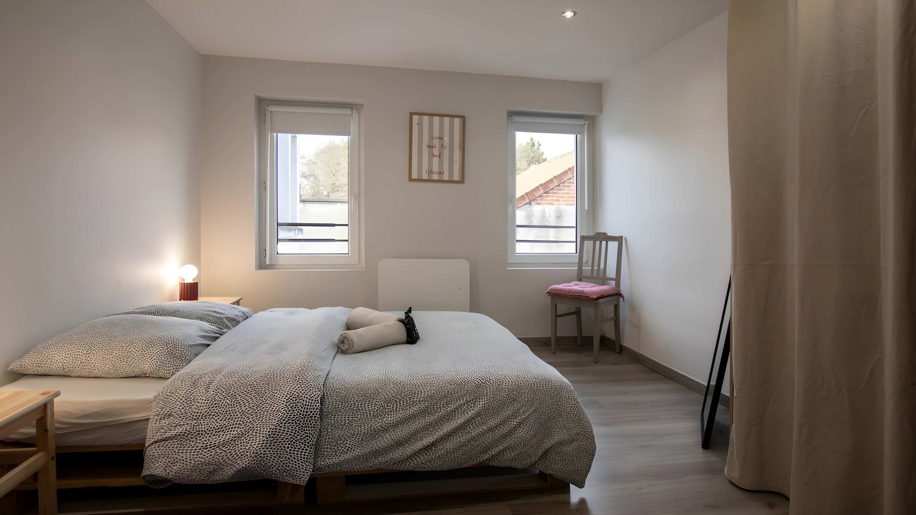 Quarto com cama, mesa de cabeceira, cadeira, duas janelas e um quadro na parede.