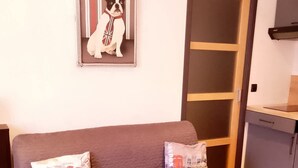 Un sofá gris con dos cojines, un cuadro enmarcado en la pared, una puerta y una mesita con una planta.