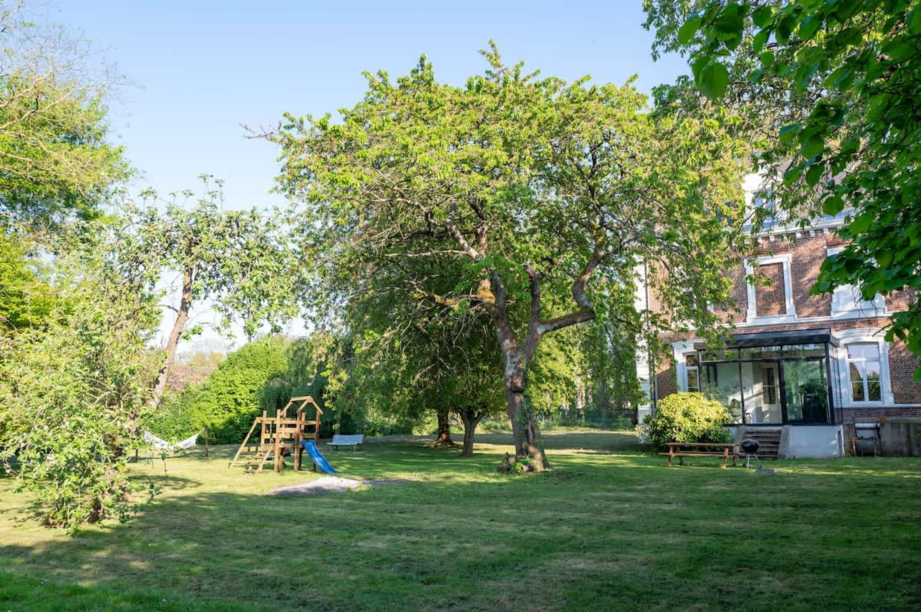 A backyard with a wooden play structure, a large tree, and a brick house with a glass conservatory.