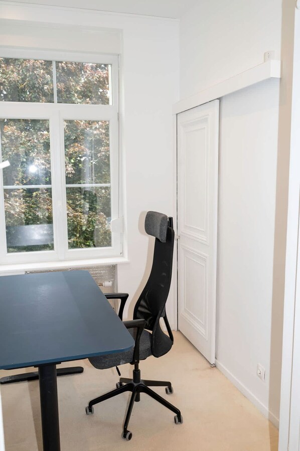 Um quarto com janela, uma mesa, uma cadeira e uma porta fechada.