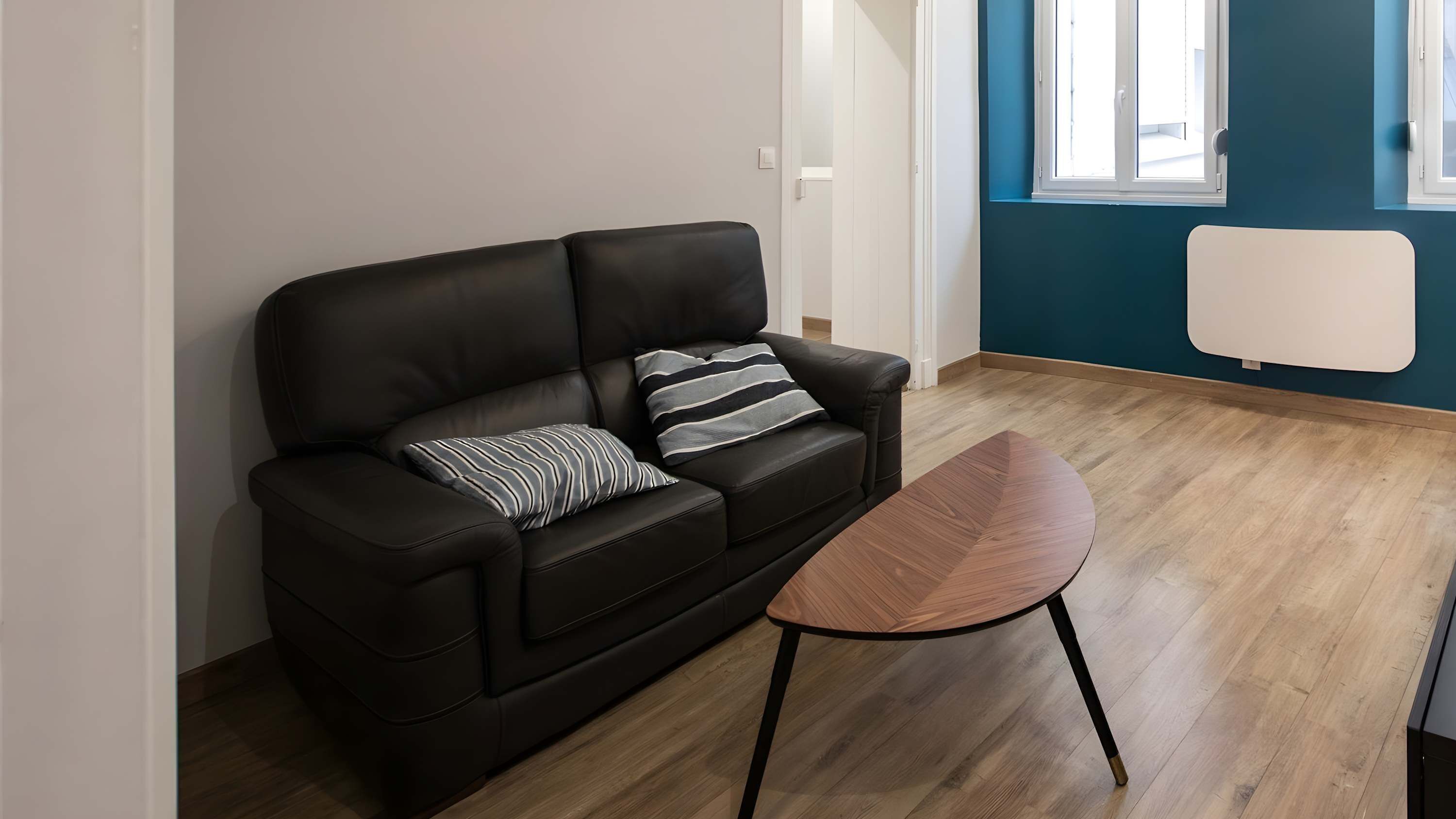 Ett modernt vardagsrum med en svart lädersoffa, ett träbord och utsikt över en blå vägg och ett fönster.