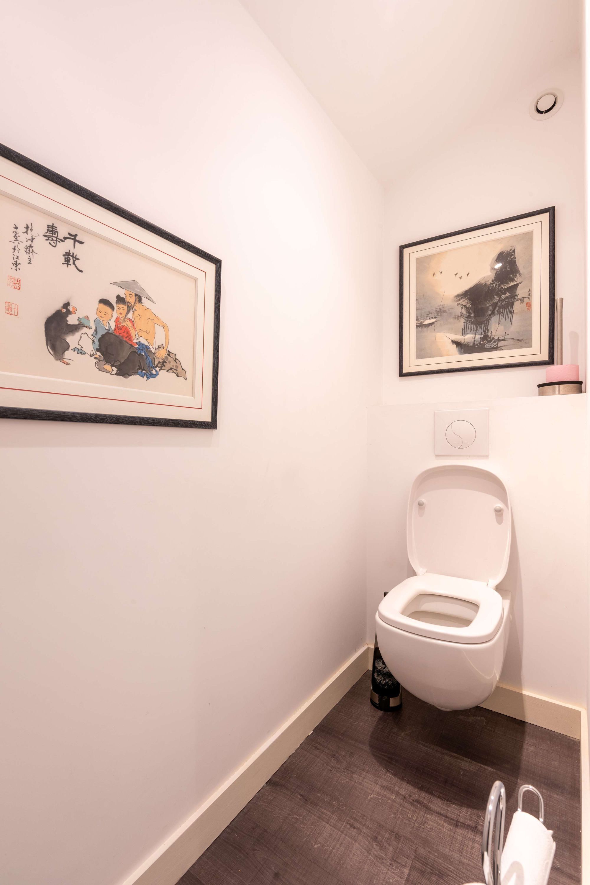 Et badeværelse med toilet, et billede hængende på væggen og en lille hylde med en flaske.
