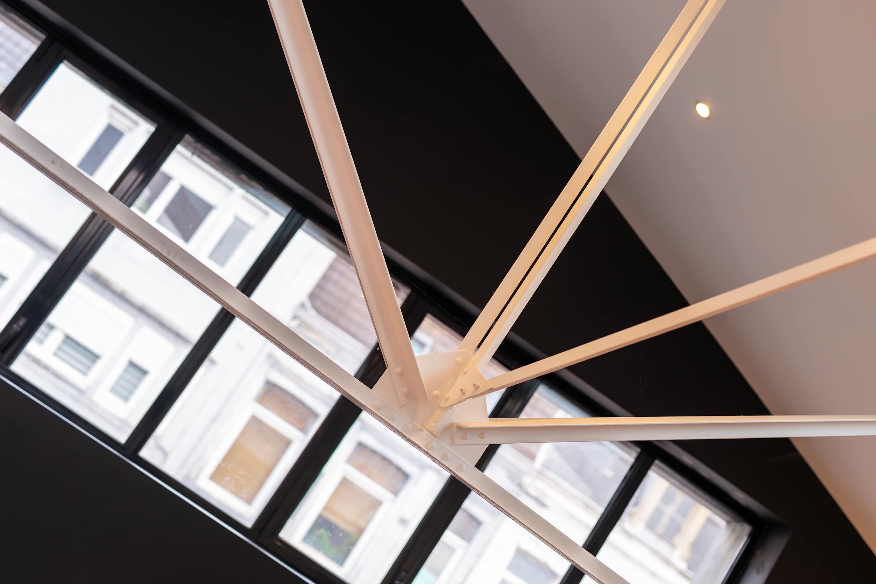 A modern ceiling structure with intersecting beams, large windows with a view of buildings, and a recessed light fixture.