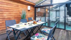 Um deck de madeira com mesa, cadeiras, um vaso de planta e uma porta de vidro que dá para uma área interna.