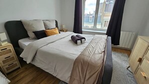 Un dormitorio ordenado con cama, mesitas de noche, una ventana con cortinas y un radiador.