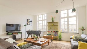 Una sala de estar con televisor, una mesa de centro, un sofá, una estantería y ventanas amplias.
