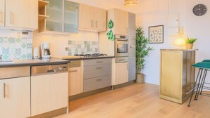 Una cocina moderna con armarios de madera clara, estufa a gas, cafetera y un comedor con una silla de metal.