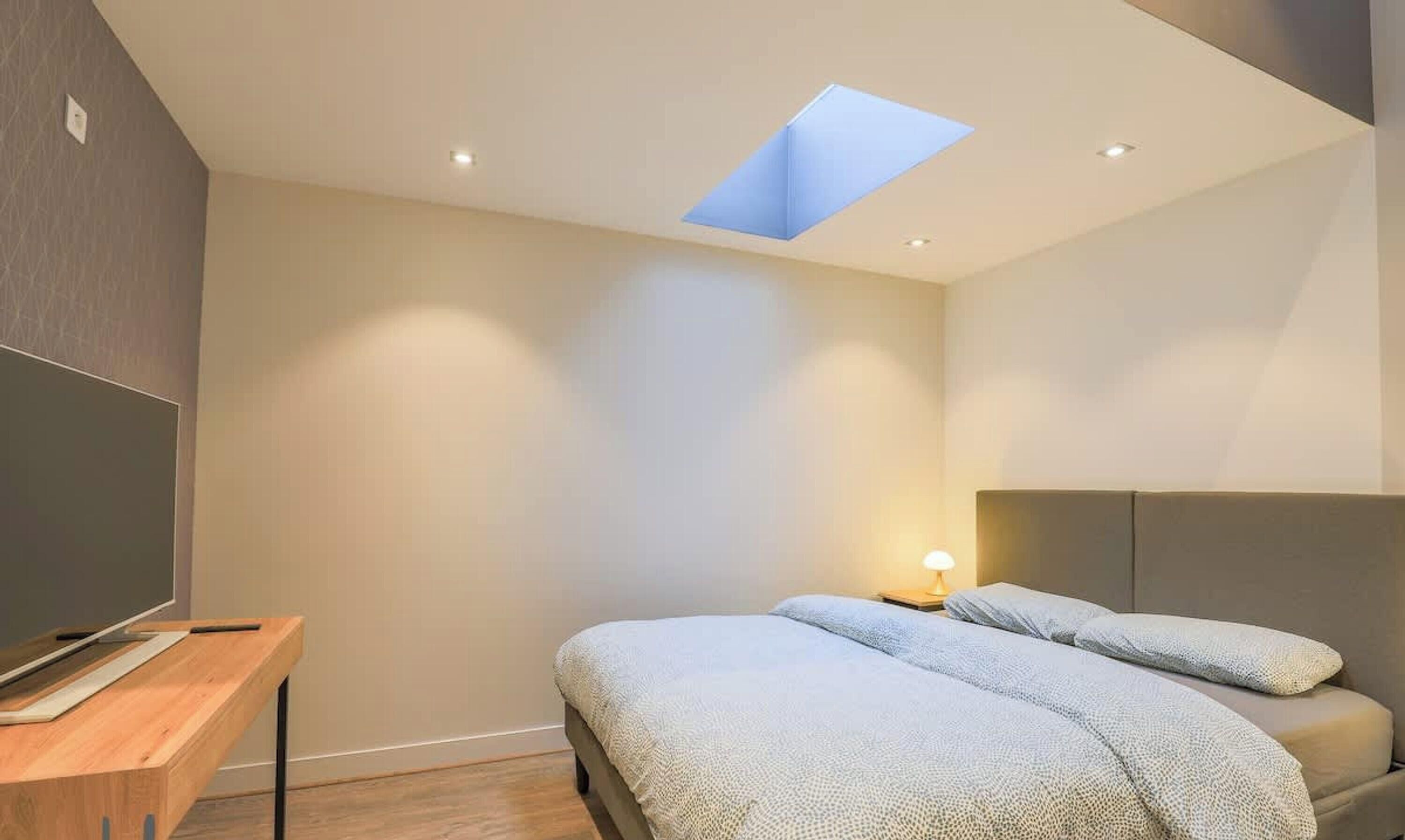 A modern bedroom with a skylight, a bed, a bedside table, and a TV.