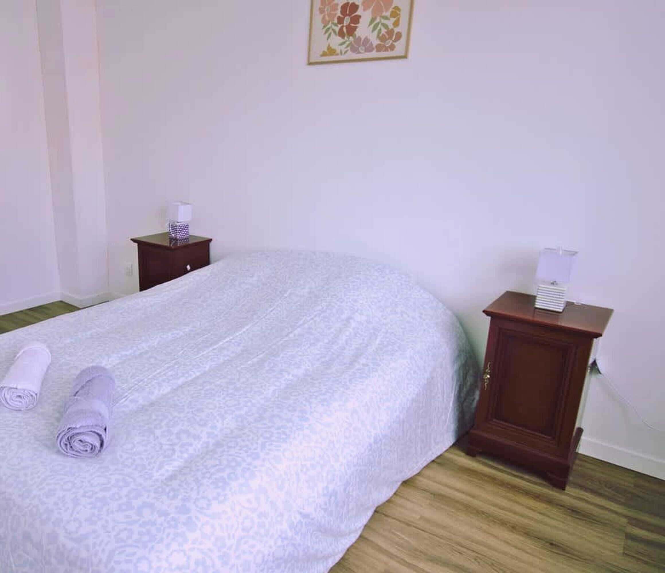 Een net opgemaakt bed met een beddekleed met patroon, twee opgerolde handdoeken, een klein houten nachtkastje met een zakdoekendoosje, en een aan de muur hangend schilderij.