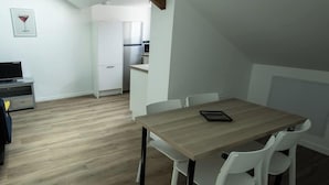 Et spiseområde med et træbord og hvide stole, et køkken i baggrunden, og et ovenlysvindue.