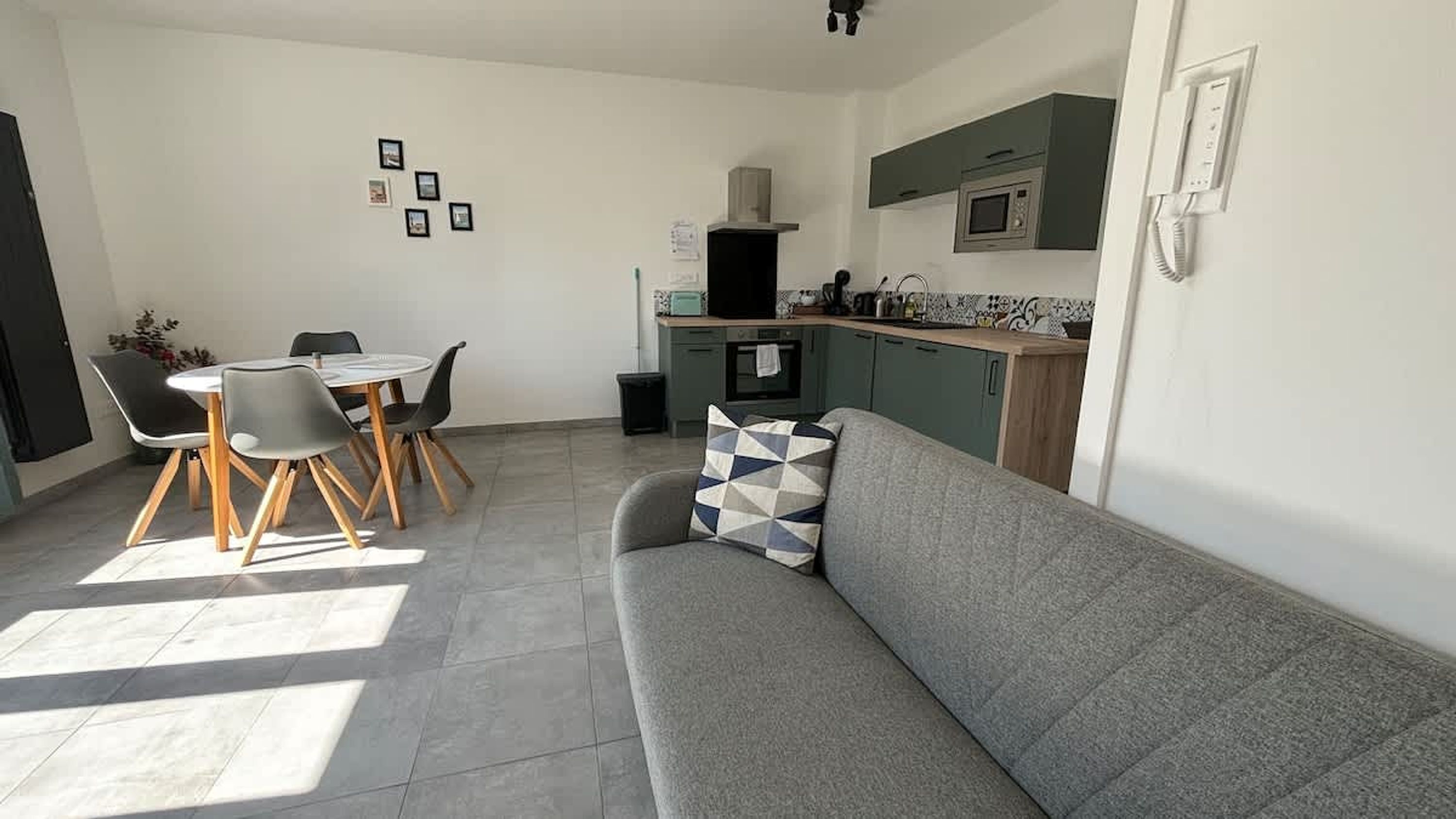 Un salon moderne avec un canapé gris, une table à manger avec des chaises, un coin cuisine équipé d’un four à micro-ondes et une petite poubelle.