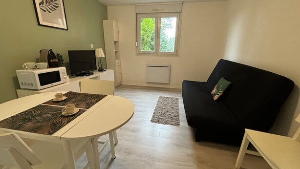 A compact living space with a dining table, a black sofa, a TV, a microwave, and a window with a view of greenery. - Studio Avec Place de Parking (Arras)