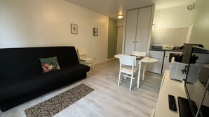 A modern living room with a black sofa, a small white table, and a kitchen area with a refrigerator and microwave. - Studio Avec Place de Parking (Arras)