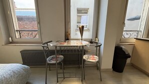 Ein kleiner Raum mit Tisch und zwei Stühlen, einem Heizkörper, einem Fenster mit Blick auf ein Gebäude und einem Mülleimer.