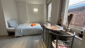 Ein kleiner, kompakter Raum mit einem Bett, einem Tisch und einem Stuhl. Ein Fenster bietet Blick auf ein Backsteingebäude.