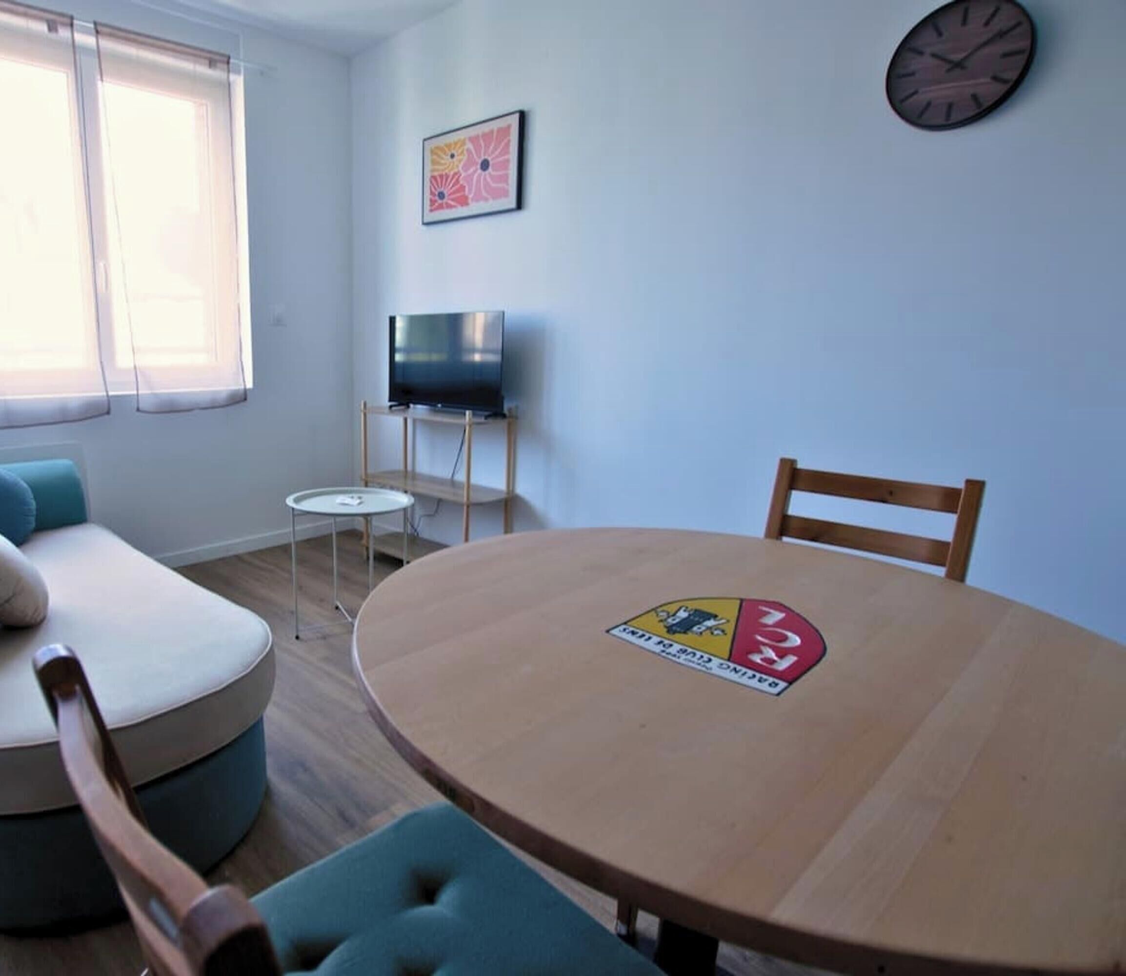 Una habitación con una mesa redonda de madera, un televisor sobre un mueble bajo, un sofá y un reloj en la pared.